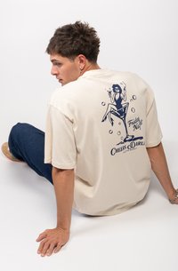 Camiseta de algodón color crema con un estampado azul marino de una mujer con un cóctel, y el texto "Friday Night Cheers. Darl" debajo del diseño.