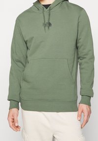 Felpa con cappuccio verde realizzata in tessuto morbido, con tasca a marsupio frontale e cappuccio con coulisse. Presenta un piccolo logo nero sul petto.