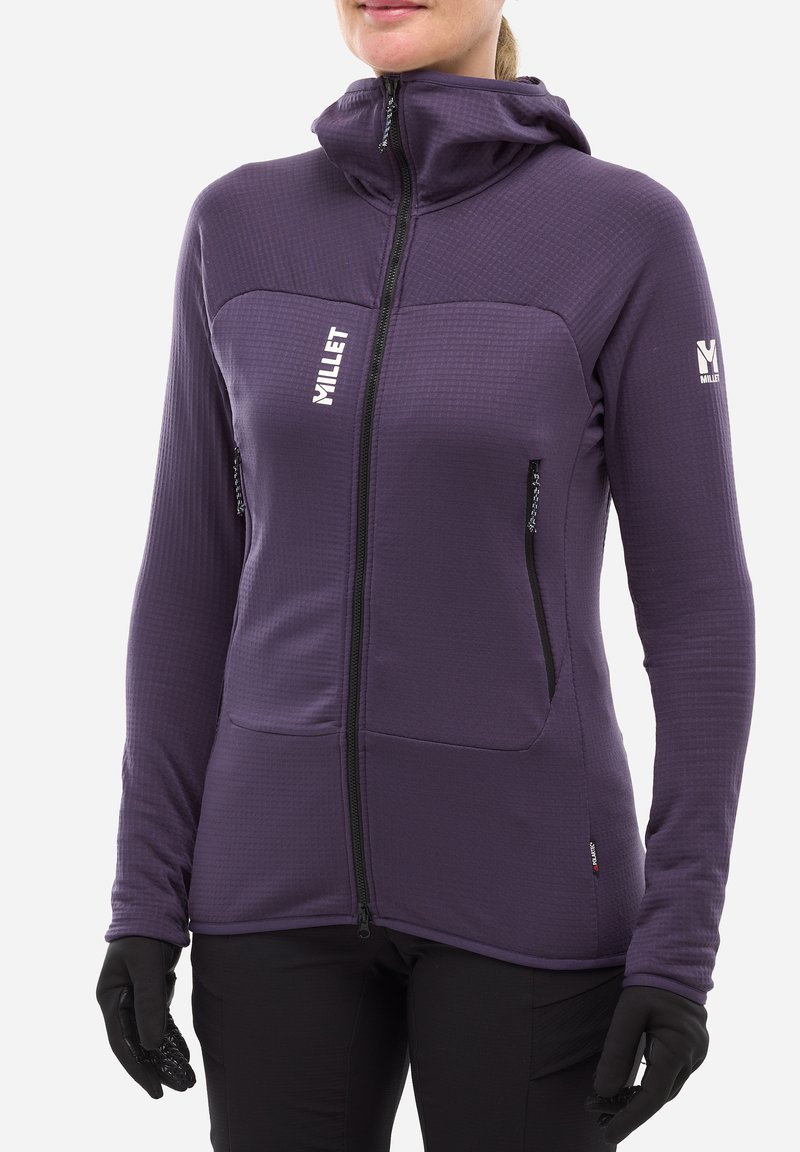 Personne portant une veste à capuche zippée violette avec des poches zippées et des gants noirs, se tenant devant un fond uni.