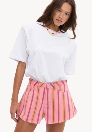 Femme portant un t-shirt blanc uni et un short rose taille haute avec des rayures verticales rouges, orange et violettes, accessoirisée d’un collier de perles volumineux.