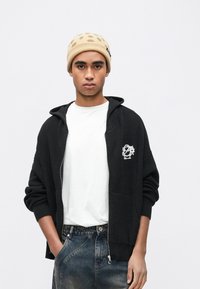 Μαύρο πλεκτό φούτερ με φερμουάρ μπροστά, διακοσμημένο με λευκό κεντημένο λογότυπο. Συνδυασμένο με απλό λευκό μπλουζάκι και φορεμένο με τζιν παντελόνι. Μπεζ σκουφί.