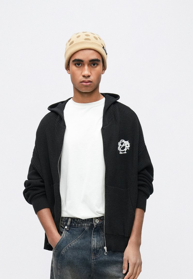 Μαύρο πλεκτό φούτερ με φερμουάρ μπροστά, διακοσμημένο με λευκό κεντημένο λογότυπο. Συνδυασμένο με απλό λευκό μπλουζάκι και φορεμένο με τζιν παντελόνι. Μπεζ σκουφί.