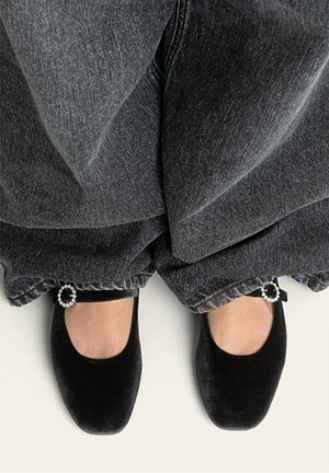 Ballerine in velluto nero con punta arrotondata e fibbia decorativa simile a una perla su pantaloni in denim scuro con orlo sfilacciato.