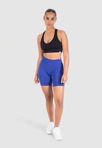 Musta urheiluliivi ja siniset korkeavyötäröiset shortsit joustavasta kankaasta, yhdistettynä valkoisiin urheilukenkiin; neutraali tausta.