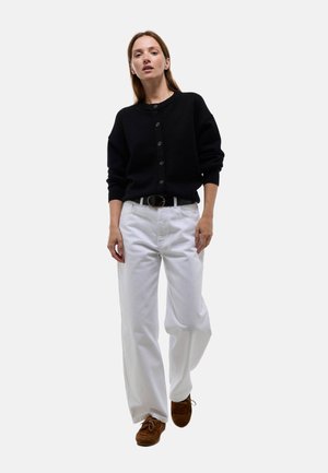 Femme portant un pull noir boutonné, un pantalon blanc à jambes larges avec une ceinture noire, et des chaussures marron à lacets, marchant vers l'avant sur un fond blanc.