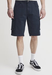 Marineblå cargo shorts med sidelommer, laget av bomullsblandet stoff. Har rett snitt, flat front og oppbrettede hem. Svarte joggesko med hvite snørebånd.