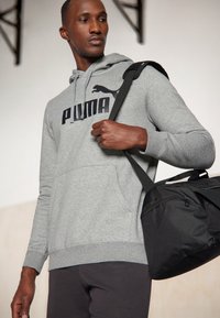 Felpa con cappuccio grigia, dotata di grande logo nero "PUMA", tasca frontale e orlo a coste, abbinata a una borsa a tracolla nera.