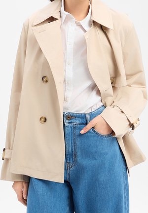 Trench-coat beige avec boutons sur une chemise blanche, associé à un jean bleu taille haute, une main dans la poche, sur un fond uni.