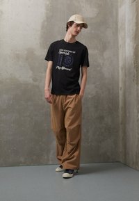 Zwarte katoenen T-shirt met witte en blauwe grafische tekst, gecombineerd met beige wijde broek en witte sneakers met blauwe details.