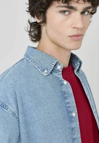 Chemise en denim bleu clair avec un col boutonné, associée à un t-shirt rouge à col rond en dessous. Le tissu semble doux avec une coupe décontractée.
