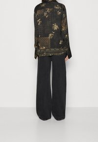 Camicia di seta nera con motivi floreali e geometrici, caratterizzata da una vestibilità rilassata e maniche lunghe, abbinata a pantaloni in denim scuro a gamba larga.