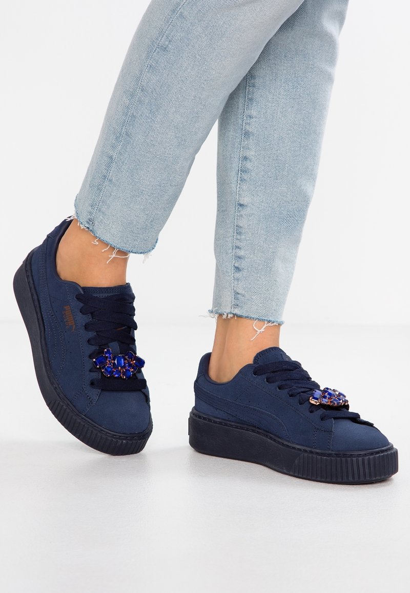 Marineblaue Plateausneaker mit schwarzen Sohlen und blauen, mit Schmucksteinen verzierten Dekorationen, getragen mit hellblauen Jeans mit fransigen Säumen vor einem weißen Hintergrund.