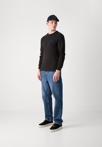 Svart langermet skjorte, denimjeans og svarte joggesko med hvite såler. Modellen har på seg en svart caps med logo. Enkel bakgrunn.