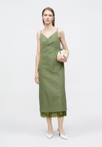 Πράσινο μίντι φόρεμα χωρίς μανίκια με σούρα μπροστά, ευθεία γραμμή και φουντωτό πράσινο τελείωμα. Συνδυάζεται με λευκά παπούτσια και μικρή τσάντα χειρός.