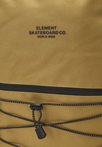 Zaino in nylon giallo con branding nero, una tasca con zip e dettagli con cordino elastico dotato di chiusura a cordoncino. Superficie testurizzata.