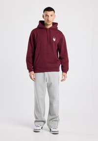Sweat à capuche bordeaux avec poche kangourou et logo, associé à un pantalon de survêtement gris avec des bandes noires sur les côtés, porté avec des baskets blanches.