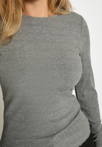 Sivi rebrasti dukserica s dugim rukavima koja sadrži male srebrne akcenta po tkanini, pružajući teksturirani izgled naspram glatke pozadine.