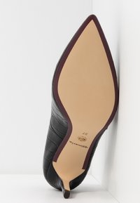 Chaussure noire à texture crocodile, bout pointu et petit talon kitten, vue de la semelle avec garniture beige et marron foncé.