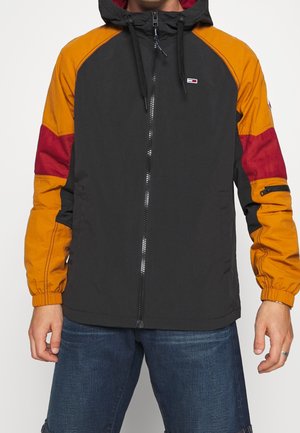 Mann trägt eine schwarze Kapuzenjacke mit orange und rot abgesetzten Ärmeln, vorderem Reißverschluss und dunkelblauen Jeans.