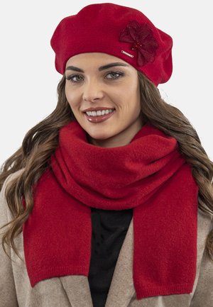 Rød uld beret med en blomsterudsmykning og et rødt tørklæde, begge med en blød tekstur, båret over en beige frakke og en sort top.