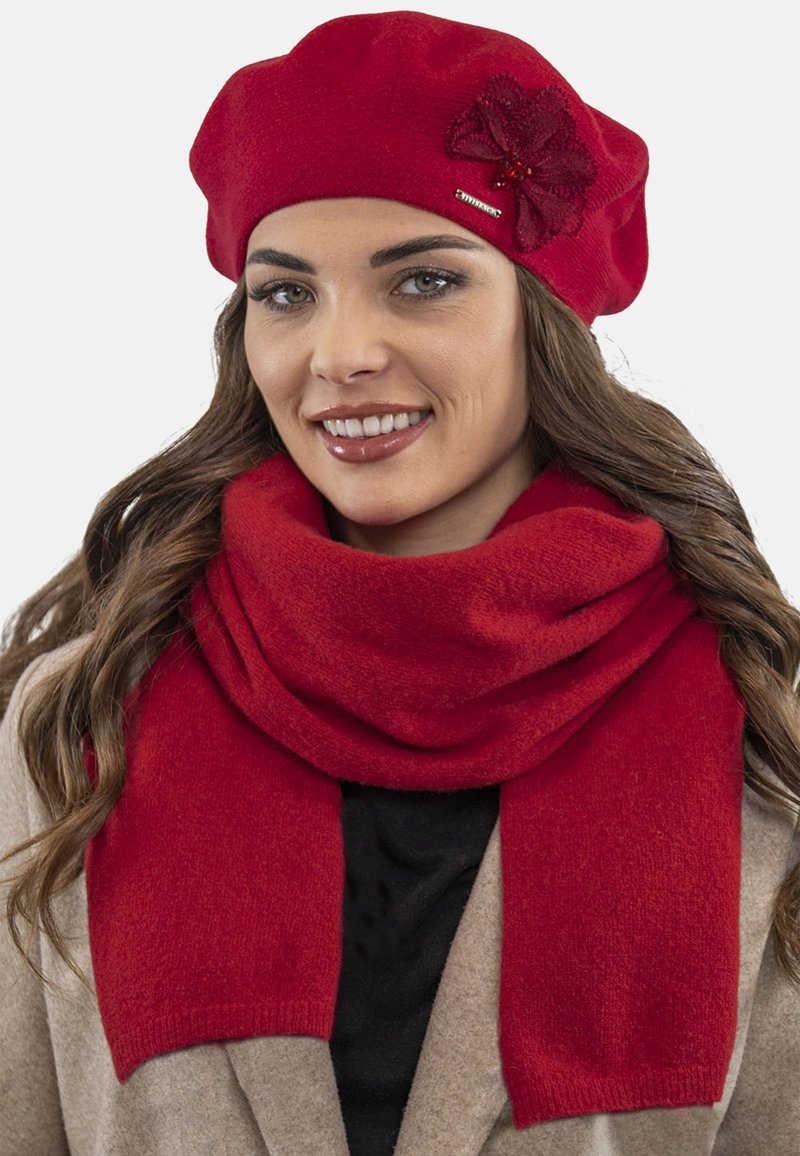Beret roșu din lână cu o aplicație florală și eșarfă roșie, ambele având o textură moale, purtate peste un palton bej și un top negru.