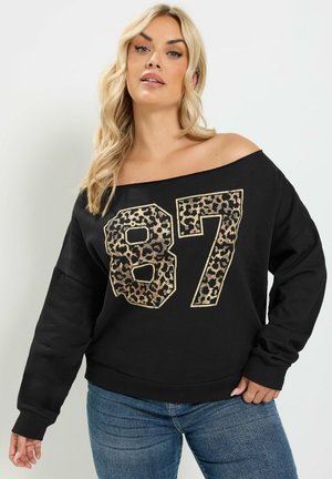 Lyshåret kvinde iført en off-shoulder sort sweatshirt med leopardprint nummer 87 og blå jeans, poserer med den ene hånd på hoften.