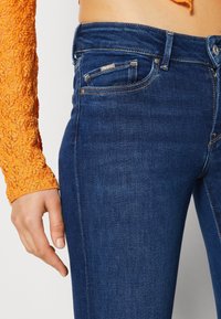 Vaqueros ajustados de mezclilla azul oscuro con una textura suave, que cuentan con un diseño clásico de cinco bolsillos y detalles de costura sutiles.