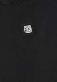 Tissu noir avec un petit badge métallique carré affichant le nom de la marque "HUGO" en lettres capitales embossées.