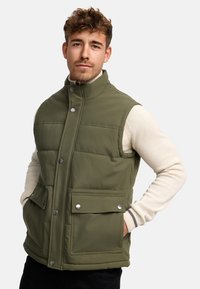 Vesta pufo de culoare verde olive, cu guler înalt, închidere cu fermoar și capse, două buzunare frontale și un design matlasat. Textură moale.