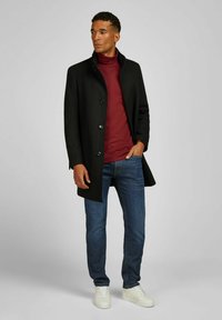 Zwarte wolenkleed met hoge kraag en knoopsluiting. Draag het over een bordeauxrode coltrui en blauwe jeans. Witte sneakers maken de outfit af.