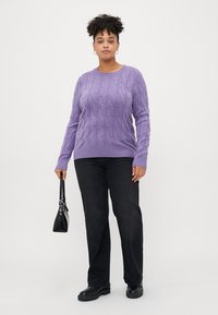 Lila Kabelstrickpullover mit rundem Halsausschnitt, kombiniert mit schwarzen, geraden Hosen und einer kleinen schwarzen Handtasche.