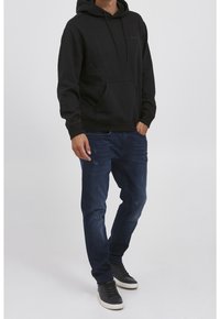 Sudadera negra con capucha y bolsillo frontal, combinada con jeans de color azul oscuro y zapatillas negras de corte bajo. Tejido suave, ajuste casual.