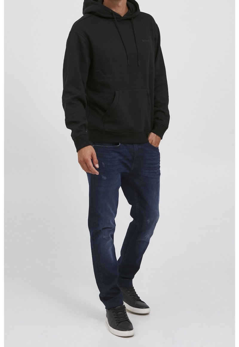 Sudadera negra con capucha y bolsillo frontal, combinada con jeans de color azul oscuro y zapatillas negras de corte bajo. Tejido suave, ajuste casual.