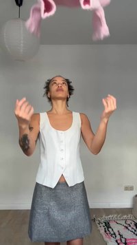 Άτομο με κοντά κατσαρά μαλλιά, λευκό αμάνικο τοπ και γκρι φούστα πετάει ένα ροζ μαντήλι προς τα επάνω σε ένα λιτά διακοσμημένο δωμάτιο.