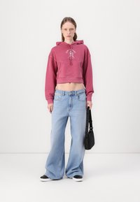 Felpa rosa corta con tasca frontale e logo; jeans larghi azzurri; borsa nera; sneakers bianche e nere.