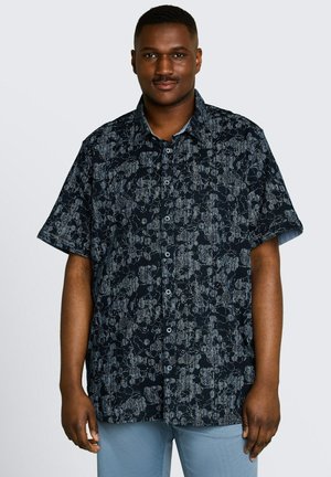 Mężczyzna stoi, ubrany w czarną krótkoturową koszulę z szarymi abstrakcyjnymi wzorami, połączoną z jasnoniebieskimi spodniami.