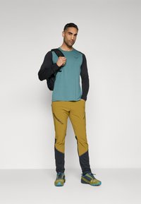 Tyrkis og sort langærmet skjorte parret med gule og sorte bukser, med et logo. Modellen holder en sort rygsæk og har robuste sneakers på.