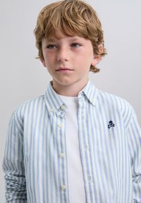 Camisa abotonada con rayas azules y verdes claras, con un logo bordado de calavera y huesos cruzados en el pecho, combinada con una camiseta blanca.