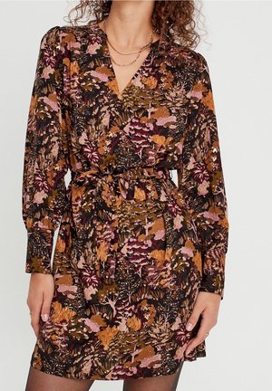 Femme portant une robe portefeuille à manches longues avec un motif floral abstrait aux tons automnaux et une ceinture nouée à la taille, accessoirisée de colliers en or superposés.