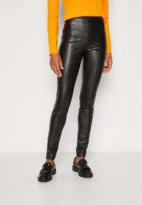 Black leather high-waisted pants with a sleek fit, smooth texture, and no visible seams. Paired with black loafers and an orange long-sleeve top.