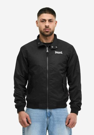 Mann trägt eine schwarze Lonsdale Zip-up Jacke mit Knopfkragen und hellblauen Jeans, steht vor weißem Hintergrund.