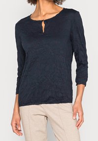 Granatowa bluzka z długim rękawem z dekoltem w kształcie łezki. Zmarszczona faktura i luźny krój. Połączona z jasnobeżowymi spodniami.