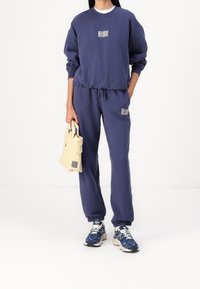 Navyblauwe sweatshirt en sweatpants set, gemaakt van katoen, met een logo op de borst en zijzakken, gecombineerd met lichte sneakers en een beige tas.