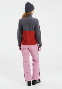 Grijze en rode fleece pullover met een hoge kraag. Gepaard met roze sneeuwbroeken met ritsen aan de zijkant en zwarte skibotten.