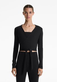 Woman wearing a black long-sleeve wrap crop top tied at the waist with high-waisted black pants, standing against a white background.