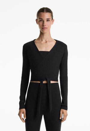Woman wearing a black long-sleeve wrap crop top tied at the waist with high-waisted black pants, standing against a white background.
