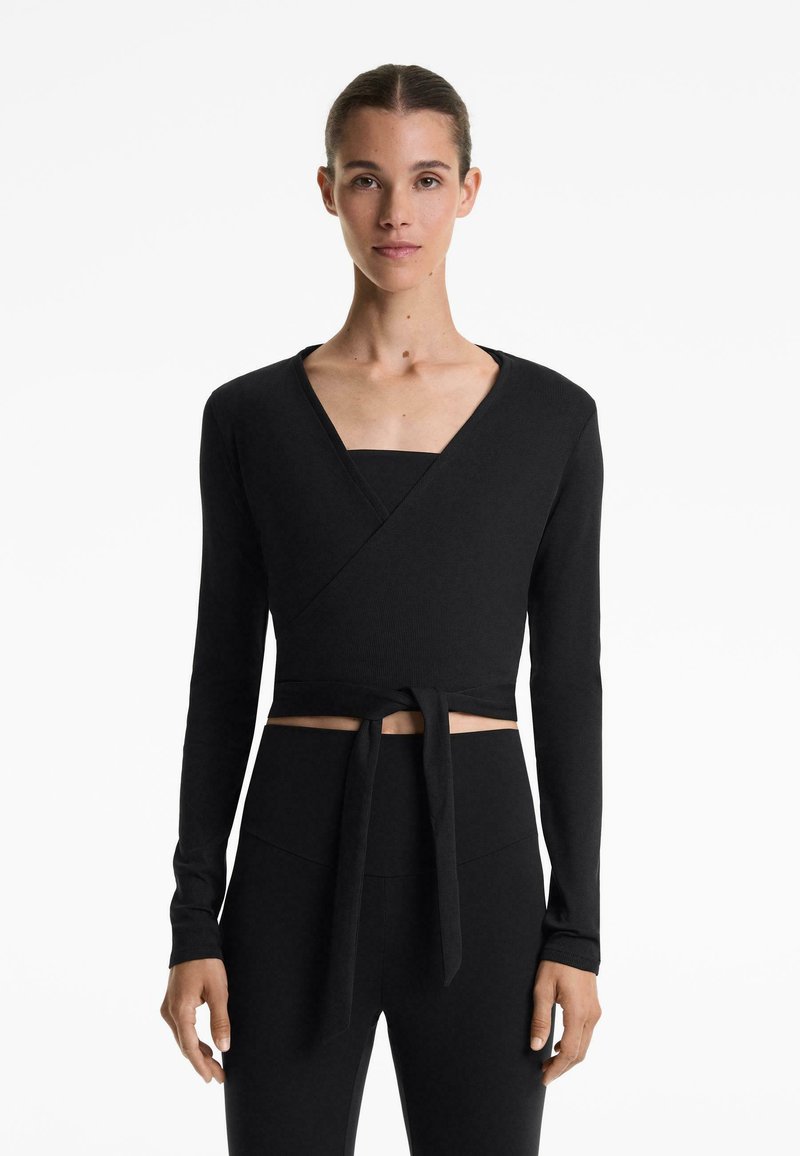 Woman wearing a black long-sleeve wrap crop top tied at the waist with high-waisted black pants, standing against a white background.