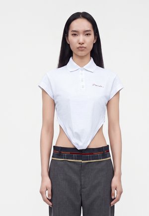Femme aux longs cheveux noirs porte un polo blanc court et un pantalon gris avec une ceinture visible et une bande rouge.