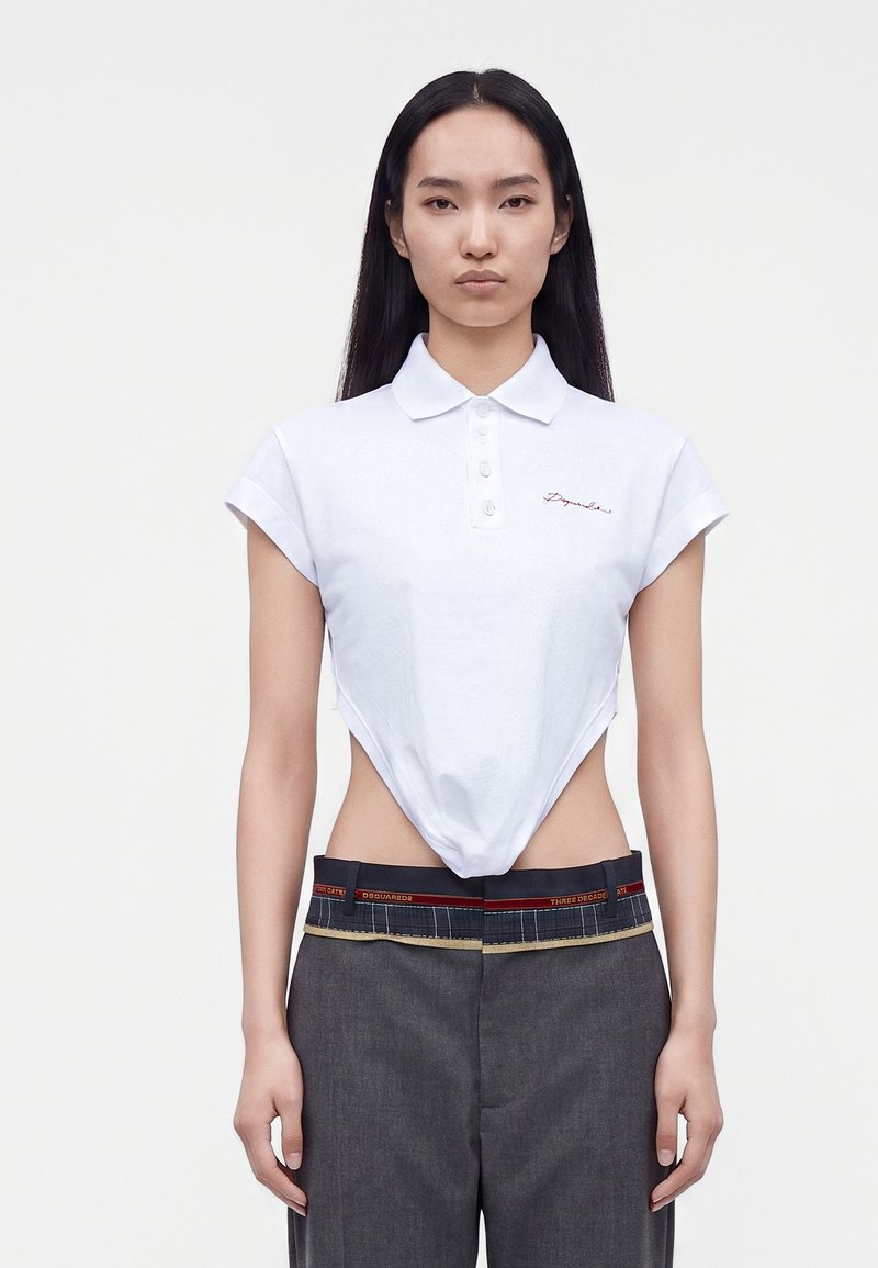 Femme aux longs cheveux noirs porte un polo blanc court et un pantalon gris avec une ceinture visible et une bande rouge.