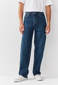 Man i mörkblå raka jeans och vita sneakers, som står mot en enkel vit bakgrund.